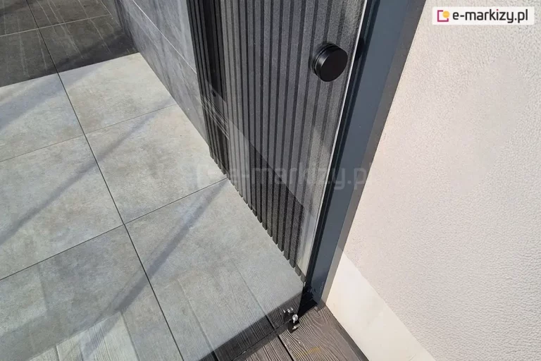 Boczne mocowanie szklanych paneli przesuwnych Miros do ściany elewacji z tynkiem strukturalnym. Na pierwszym panelu szklanym widoczna okrągła czarna klamka oraz stoper, ograniczający możliwość otwarcia drzwi. W tle za szyba widoczny taras wyłożony płytami gresowymi.