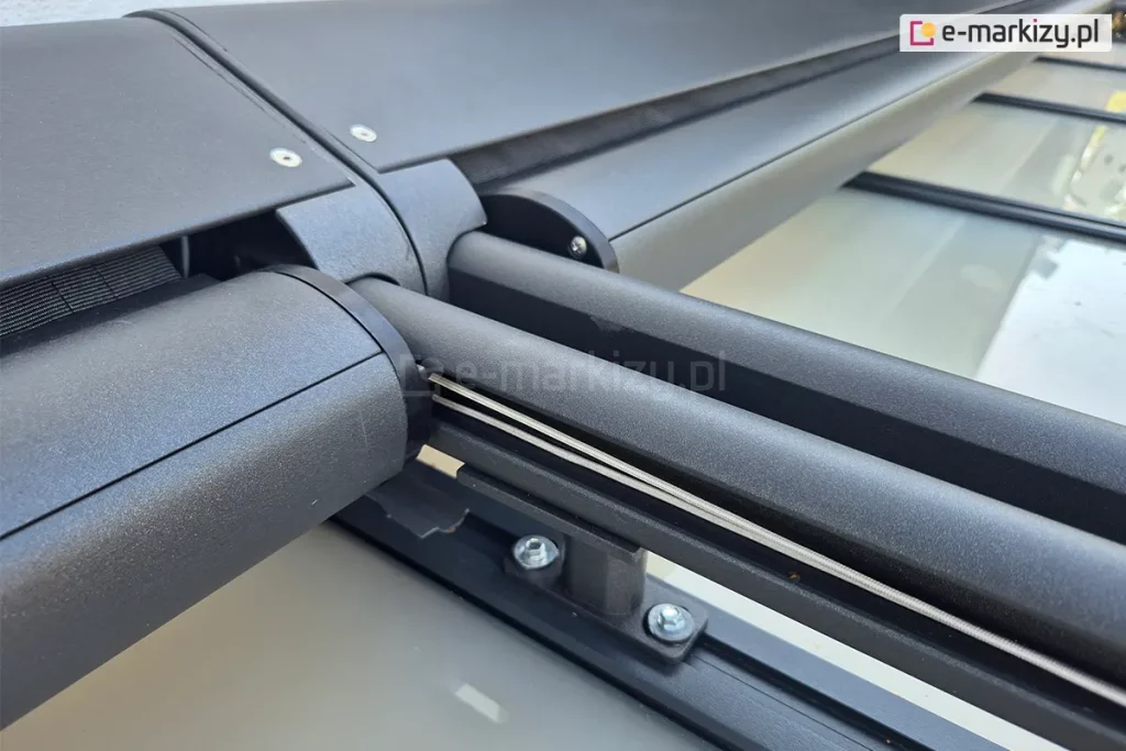 Zbliżenie uchwytu modułowego systemu veranda Selt, w którym zamocowane są dwie prowadnice oraz kasety w kolorze antracytu. W jednej z prowadnic widoczna linka odpowiadająca za prawidłowe naprężenie tkaniny rolety poziomej.