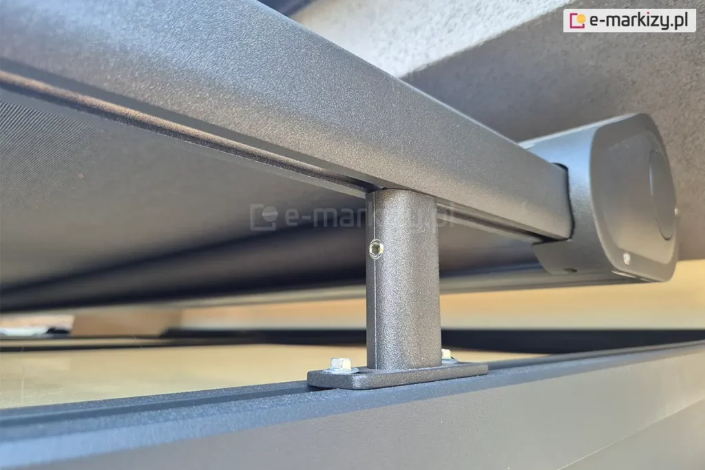 Zdjęcie przedstawia techniczne zbliżenie na detale montażowe systemu veranda Aluprof. Widoczny jest aluminiowy profil prowadzący oraz zaokrąglona zaślepka kasety, lakierowane proszkowo na kolor ciemnoantracytowy o drobnoziarnistej strukturze. Konstrukcja wspiera się na pionowym, krótkim wsporniku (stopie montażowej), który jest przytwierdzony do dolnego profilu za pomocą dwóch stalowych śrub. W górnej części kadru widoczna jest napięta tkanina w kolorze grafitowym. Elementy są zamontowane w bliskiej odległości od jasnej, tynkowanej elewacji budynku, widocznej w tle. Zdjęcie kładzie nacisk na solidność połączeń mechanicznych oraz estetyczne wykończenie komponentów systemu osłonowego.