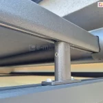 Zdjęcie przedstawia techniczne zbliżenie na detale montażowe systemu veranda Aluprof. Widoczny jest aluminiowy profil prowadzący oraz zaokrąglona zaślepka kasety, lakierowane proszkowo na kolor ciemnoantracytowy o drobnoziarnistej strukturze. Konstrukcja wspiera się na pionowym, krótkim wsporniku (stopie montażowej), który jest przytwierdzony do dolnego profilu za pomocą dwóch stalowych śrub. W górnej części kadru widoczna jest napięta tkanina w kolorze grafitowym. Elementy są zamontowane w bliskiej odległości od jasnej, tynkowanej elewacji budynku, widocznej w tle. Zdjęcie kładzie nacisk na solidność połączeń mechanicznych oraz estetyczne wykończenie komponentów systemu osłonowego.