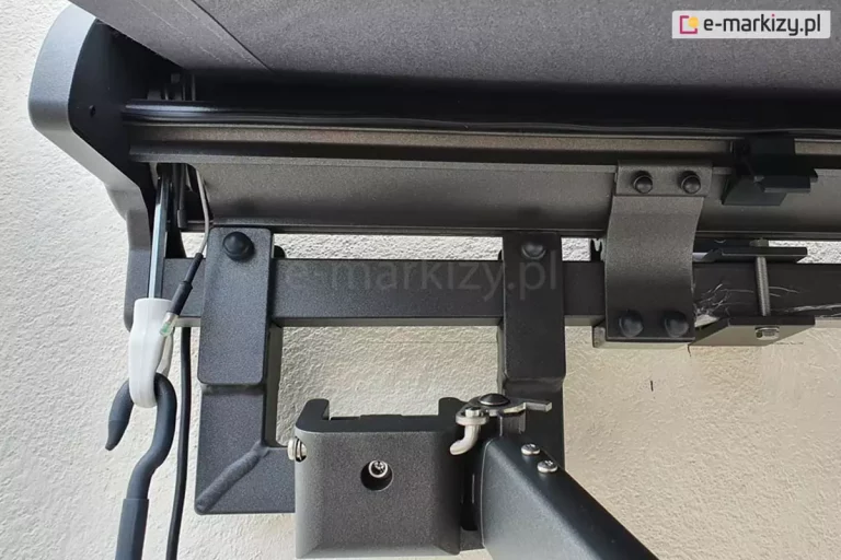 Zbliżenie belki kwadratowej markizy Palladio Selt w kolorze srebrzysty grafit z widocznym mocowaniem ramienia krzyżowego oraz mocowania kasety i uchwytu ściennego po prawej i przekładnią korbową do sterowania ręcznego z lewej. W tle biała ściana z tynkiem strukturalnym .