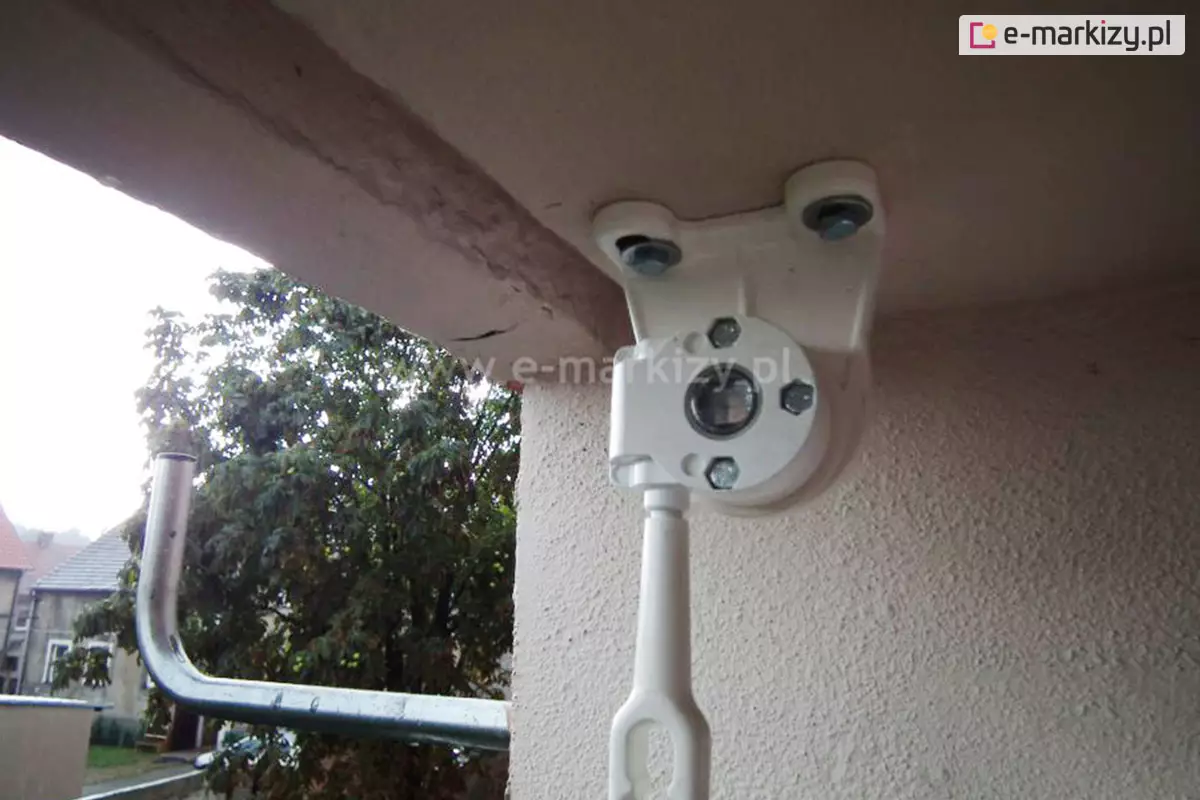 Zdjęcie prezentuje zbliżenie na biały uchwyt montażowy z zainstalowaną przekładnią oczkową, widoczny z perspektywy od dołu. Mechanizm jest przykręcony do sufitu wnęki balkonowej za pomocą dwóch widocznych śrub z podkładkami. Centralnym punktem jest okrągła obudowa przekładni, w której osadzony jest otwór na trzpień rury nawojowej markizy. Pod obudową znajduje się pionowo skierowane białe oczko do korby ręcznej. Po lewej stronie kadru, na dalszym planie, widoczny jest fragment wygiętej, srebrnej pustej rury montażowej do anteny satelitarnej. W tle za barierką rozpościera się widok na osiedle – widoczne są zielone korony drzew oraz fragmenty budynków mieszkalnych o jasnych elewacjach i spadzistych dachach. Całość kompozycji osadzona jest w kontekście zewnętrznym, na balkonie, przy jasnej ścianie o chropowatej fakturze tynku. Światło dzienne dobrze doświetla detale konstrukcyjne przekładni oraz sposób jej zamocowania do elementu nośnego.