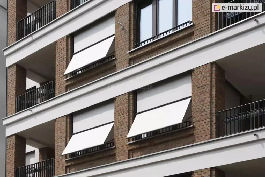 Zdjęcie prezentuje fragment wielopiętrowego budynku o nowoczesnej architekturze, na którego elewacji zainstalowano systemy markizolet. Fasada wykonana jest z brązowej cegły klinkierowej, poprzeczkowanej białymi, poziomymi pasami konstrukcyjnymi oddzielającymi kondygnacje. W pionowych rzędach okien i loggii widoczne są osłony przeciwsłoneczne z białą, gładką tkaniną. Trzy markizolety są w pełni rozwinięte i posiadają odchyloną dolną część, która tworzy skośny daszek u dołu otworu okiennego, natomiast jedna osłona w górnej części zdjęcia pozostaje całkowicie zwinięta w kasecie. Konstrukcja ramion oraz prowadnic bocznych została wykonana w ciemnym kolorze, kontrastującym z jasnym poszyciem materiału. Po lewej stronie oraz w narożnikach widoczne są balkony z ażurowymi, ciemnymi balustradami, które dopełniają techniczną estetykę budynku.