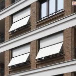 Zdjęcie prezentuje fragment wielopiętrowego budynku o nowoczesnej architekturze, na którego elewacji zainstalowano systemy markizolet. Fasada wykonana jest z brązowej cegły klinkierowej, poprzeczkowanej białymi, poziomymi pasami konstrukcyjnymi oddzielającymi kondygnacje. W pionowych rzędach okien i loggii widoczne są osłony przeciwsłoneczne z białą, gładką tkaniną. Trzy markizolety są w pełni rozwinięte i posiadają odchyloną dolną część, która tworzy skośny daszek u dołu otworu okiennego, natomiast jedna osłona w górnej części zdjęcia pozostaje całkowicie zwinięta w kasecie. Konstrukcja ramion oraz prowadnic bocznych została wykonana w ciemnym kolorze, kontrastującym z jasnym poszyciem materiału. Po lewej stronie oraz w narożnikach widoczne są balkony z ażurowymi, ciemnymi balustradami, które dopełniają techniczną estetykę budynku.