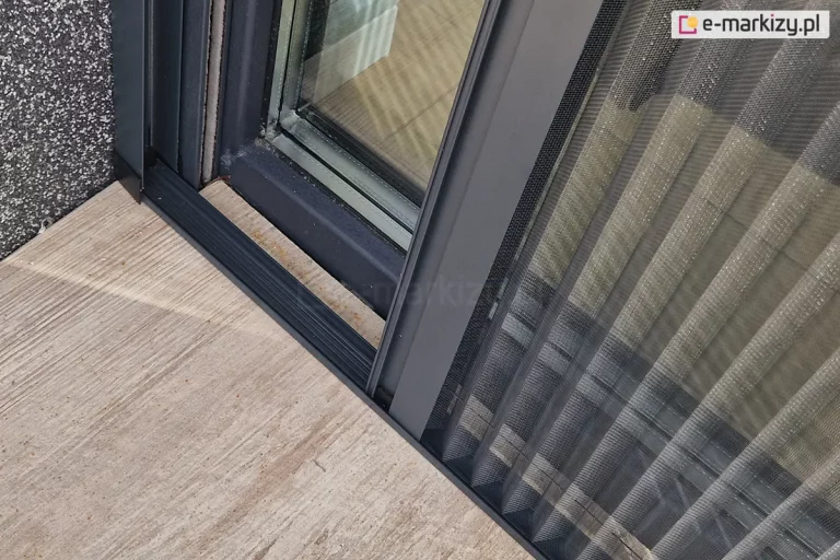 Zbliżenie prowadnicy dolnej moskitiery, w której widoczne są sznurki odpowiadające za rozwijanie i składanie plisowanej siatki. Próg w kolorze konstrukcji moskitiery zamocowany został do podłogi tarasu wyłożonego drewnopodobnymi płytkami w lekkim oddaleniu od ramy okna z uwagi na występujące przy nim zintegrowane rolety zewnętrzne.