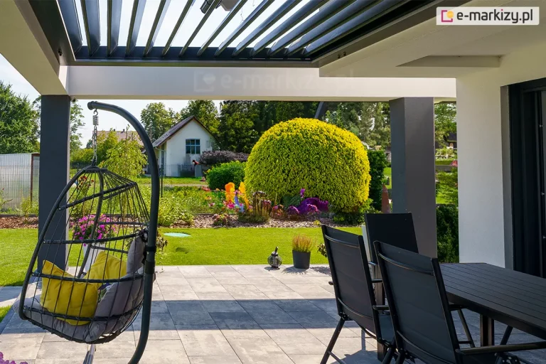widok z wnętrza nowoczesnej pergoli SB400R na ogród, skupiając się na funkcjonalności ruchomych lameli dachowych. Aluminiowe lamele w kolorze antracytowym są ustawione w pozycji uchylonej pod kątem około 45 stopni, co pozwala na jednoczesną wentylację przestrzeni oraz naturalne doświetlenie tarasu przy zachowaniu ochrony przed bezpośrednim słońcem. Konstrukcja dachu opiera się na białych, szerokich belkach poprzecznych i ciemnoszarych słupach nośnych. Pod zadaszeniem znajduje się strefa wypoczynkowa z nowoczesnymi meblami: czarnym stołem z krzesłami oraz wiszącym fotelem typu "kokon" z szarymi poduszkami, zamontowanym na stabilnym stelażu. Posadzka tarasu została wykonana z dużych, jasnoszarych płyt betonowych. W tle rozpościera się widok na zadbany trawnik, formowane krzewy ozdobne o intensywnie żółtych i zielonych barwach oraz fragmenty sąsiedniej zabudowy pod pogodnym, jasnym niebem.