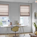 Jasne i inspirujące wnętrze domowego biura lub pracowni artystycznej, w którym dwa wysokie, pionowe okna zostały wyposażone w stylowe rolety rzymskie. Przesłony wykonane są z tkaniny o grubym splocie w naturalnym kolorze ciepłego beżu lub jasnego brązu. Rolety rzymskie są podciągnięte do połowy wysokości okien, tworząc w dolnej części miękkie, estetyczne fałdy, co pozwala na swobodny dopływ światła dziennego do miejsca pracy, jednocześnie chroniąc przed bezpośrednim nasłonecznieniem. Pomieszczenie ma białe ściany i jasną podłogę ułożoną w klasyczną jodełkę. Pod oknami ustawiono szerokie, białe biurko na smukłych nogach typu kozioł, przy którym stoi nowoczesny fotel biurowy w kolorze miodowym na metalowej podstawie. Na blacie biurka panuje artystyczny nieład: widać tam grafikę w ramce, kubek, przybory do pisania oraz niebieską lampkę biurkową o geometrycznym kształcie. Po prawej stronie biurka, w rogu pokoju, znajduje się strefa relaksu z niskim, designerskim fotelem w kolorze brudnego różu, ozdobionym musztardową poduszką. Obok fotela na małym stoliku stoi donica z wysoką sansewierią, a na ścianie powyżej wiszą dwa pionowe obrazy przedstawiające naturę. Po lewej stronie pokoju, przy krawędzi okna, pionowo ustawiono bele materiałów w odcieniach fioletu i beżu. Przez okna widać fragmenty miejskiej zabudowy z ceglaną elewacją. Całość aranżacji jest lekka i nowoczesna, a beżowe rolety rzymskie idealnie dopełniają przytulny, a zarazem profesjonalny charakter wnętrza.