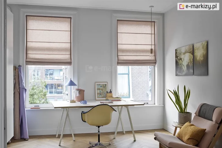 Jasne i inspirujące wnętrze domowego biura lub pracowni artystycznej, w którym dwa wysokie, pionowe okna zostały wyposażone w stylowe rolety rzymskie. Przesłony wykonane są z tkaniny o grubym splocie w naturalnym kolorze ciepłego beżu lub jasnego brązu. Rolety rzymskie są podciągnięte do połowy wysokości okien, tworząc w dolnej części miękkie, estetyczne fałdy, co pozwala na swobodny dopływ światła dziennego do miejsca pracy, jednocześnie chroniąc przed bezpośrednim nasłonecznieniem. Pomieszczenie ma białe ściany i jasną podłogę ułożoną w klasyczną jodełkę. Pod oknami ustawiono szerokie, białe biurko na smukłych nogach typu kozioł, przy którym stoi nowoczesny fotel biurowy w kolorze miodowym na metalowej podstawie. Na blacie biurka panuje artystyczny nieład: widać tam grafikę w ramce, kubek, przybory do pisania oraz niebieską lampkę biurkową o geometrycznym kształcie. Po prawej stronie biurka, w rogu pokoju, znajduje się strefa relaksu z niskim, designerskim fotelem w kolorze brudnego różu, ozdobionym musztardową poduszką. Obok fotela na małym stoliku stoi donica z wysoką sansewierią, a na ścianie powyżej wiszą dwa pionowe obrazy przedstawiające naturę. Po lewej stronie pokoju, przy krawędzi okna, pionowo ustawiono bele materiałów w odcieniach fioletu i beżu. Przez okna widać fragmenty miejskiej zabudowy z ceglaną elewacją. Całość aranżacji jest lekka i nowoczesna, a beżowe rolety rzymskie idealnie dopełniają przytulny, a zarazem profesjonalny charakter wnętrza.