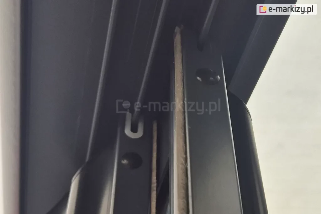 Zdjęcie przedstawia szczegółowe zbliżenie na górne wykończenie pionowych profili aluminiowych systemu przesuwnego Sotro, zamontowanych w wielotorowej prowadnicy sufitowej. Na ujęciu widoczne są dwa równoległe panele, których ramy w kolorze matowego antracytu są wyposażone w zintegrowane systemy uszczelnień. W górnej części profili znajdują się białe uszczelki ślizgowe w kształcie litery U, które obejmują prowadnice, zapewniając stabilne i ciche przesuwanie skrzydeł. Dodatkowo wzdłuż pionowych krawędzi profili przytwierdzone są gęste uszczelki szczotkowe w kolorze szarym, które wypełniają przestrzeń między panelami, chroniąc wnętrze zabudowy przed wiatrem, kurzem oraz insektami. Perspektywa od dołu ukazuje również okrągłe otwory technologiczne w bocznych ściankach profili, służące do montażu i regulacji komponentów wewnętrznych systemu.