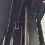 Zdjęcie przedstawia szczegółowe zbliżenie na górne wykończenie pionowych profili aluminiowych systemu przesuwnego Sotro, zamontowanych w wielotorowej prowadnicy sufitowej. Na ujęciu widoczne są dwa równoległe panele, których ramy w kolorze matowego antracytu są wyposażone w zintegrowane systemy uszczelnień. W górnej części profili znajdują się białe uszczelki ślizgowe w kształcie litery U, które obejmują prowadnice, zapewniając stabilne i ciche przesuwanie skrzydeł. Dodatkowo wzdłuż pionowych krawędzi profili przytwierdzone są gęste uszczelki szczotkowe w kolorze szarym, które wypełniają przestrzeń między panelami, chroniąc wnętrze zabudowy przed wiatrem, kurzem oraz insektami. Perspektywa od dołu ukazuje również okrągłe otwory technologiczne w bocznych ściankach profili, służące do montażu i regulacji komponentów wewnętrznych systemu.