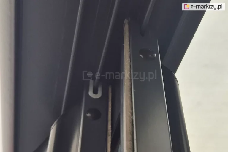 Zdjęcie przedstawia szczegółowe zbliżenie na górne wykończenie pionowych profili aluminiowych systemu przesuwnego Sotro, zamontowanych w wielotorowej prowadnicy sufitowej. Na ujęciu widoczne są dwa równoległe panele, których ramy w kolorze matowego antracytu są wyposażone w zintegrowane systemy uszczelnień. W górnej części profili znajdują się białe uszczelki ślizgowe w kształcie litery U, które obejmują prowadnice, zapewniając stabilne i ciche przesuwanie skrzydeł. Dodatkowo wzdłuż pionowych krawędzi profili przytwierdzone są gęste uszczelki szczotkowe w kolorze szarym, które wypełniają przestrzeń między panelami, chroniąc wnętrze zabudowy przed wiatrem, kurzem oraz insektami. Perspektywa od dołu ukazuje również okrągłe otwory technologiczne w bocznych ściankach profili, służące do montażu i regulacji komponentów wewnętrznych systemu.