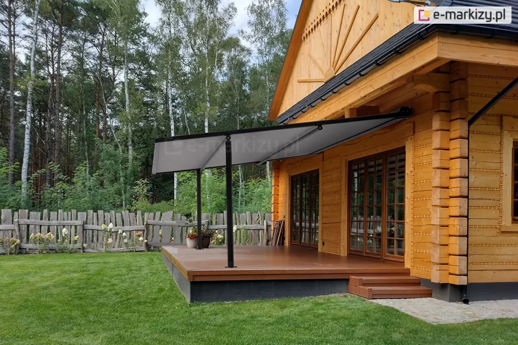 Lekka pergola tarasowa przyścienna Vera-Sola o minimalistycznej konstrukcji aluminiowej w kolorze czarnym, zamontowana do elewacji klasycznego domu z bali drewnianych. Zadaszenie stanowi jednolite, gładkie poszycie tkaninowe w kolorze szarym, które jest całkowicie rozłożone pod skosem i wsparte na dwóch smukłych, czarnych profilach pionowych przymocowanych do krawędzi tarasu. Taras wykonany jest z brązowych desek kompozytowych i osadzony na ciemnej podmurówce, a prowadzą na niego niskie, drewniane schodki. Na tarasie, obok słupa konstrukcyjnego, ustawiono dwie donice z kwitnącymi roślinami. W tle widoczny jest wysoki, drewniany płot o nieregularnym wykończeniu oraz gęsty las iglasty pod lekko zachmurzonym niebem. Zdjęcie wykonane w świetle dziennym, prezentuje estetyczny kontrast między nowoczesnym systemem zadaszenia a tradycyjną architekturą drewnianą budynku.