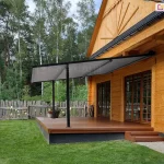 Lekka pergola tarasowa przyścienna Vera-Sola o minimalistycznej konstrukcji aluminiowej w kolorze czarnym, zamontowana do elewacji klasycznego domu z bali drewnianych. Zadaszenie stanowi jednolite, gładkie poszycie tkaninowe w kolorze szarym, które jest całkowicie rozłożone pod skosem i wsparte na dwóch smukłych, czarnych profilach pionowych przymocowanych do krawędzi tarasu. Taras wykonany jest z brązowych desek kompozytowych i osadzony na ciemnej podmurówce, a prowadzą na niego niskie, drewniane schodki. Na tarasie, obok słupa konstrukcyjnego, ustawiono dwie donice z kwitnącymi roślinami. W tle widoczny jest wysoki, drewniany płot o nieregularnym wykończeniu oraz gęsty las iglasty pod lekko zachmurzonym niebem. Zdjęcie wykonane w świetle dziennym, prezentuje estetyczny kontrast między nowoczesnym systemem zadaszenia a tradycyjną architekturą drewnianą budynku.