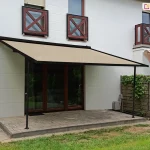 Nowoczesna pergola tarasowa przyścienna Vera-Sola o lekkiej konstrukcji aluminiowej w kolorze czarnym, zamontowana do białej elewacji piętrowego domu jednorodzinnego. Zadaszenie stanowi gładkie poszycie tkaninowe w kolorze beżowym, które jest całkowicie rozłożone pod kątem, wspierając się na dwóch smukłych profilach pionowych zamocowanych do podłoża. Pergola osłania taras wyłożony szarymi, prostokątnymi płytami chodnikowymi, na który prowadzą przeszklone drzwi tarasowe. Po prawej stronie konstrukcji znajduje się drewniane ogrodzenie panelowe częściowo porośnięte zielonymi pnączami oraz donice z roślinnością, a w tle widoczny jest fragment sąsiedniego balkonu z brązową barierką i wysuniętą markizą w czerwono-beżowe pasy. Całość znajduje się na tle zadbanego trawnika w pełnym świetle dziennym, prezentując minimalistyczne podejście do ochrony przeciwsłonecznej strefy wypoczynkowej.