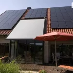 Dom jednorodzinny z dachem pokrytym czerwoną dachówką ceramiczną, na którym zamontowano instalację fotowoltaiczną oraz zewnętrzną markizę dachową nad przeszklonym ogrodem zimowym. Panele fotowoltaiczne w kolorze czarnym rozmieszczone są w trzech sekcjach na połaci dachu. Centralna część dachu nad wykuszem wyposażona jest w jasnoszarą roletę veranda V-ziiip, która całkowicie zasłania szklane zadaszenie tarasu. Przed domem znajduje się drewniany taras z meblami ogrodowymi: okrągłym stołem, krzesłami z pasiastymi poduszkami, leżakami oraz rozłożonym czerwonym parasolem przeciwsłonecznym. Elewacja budynku wykonana z jasnej cegły, otoczona gęstą roślinnością ogrodową.