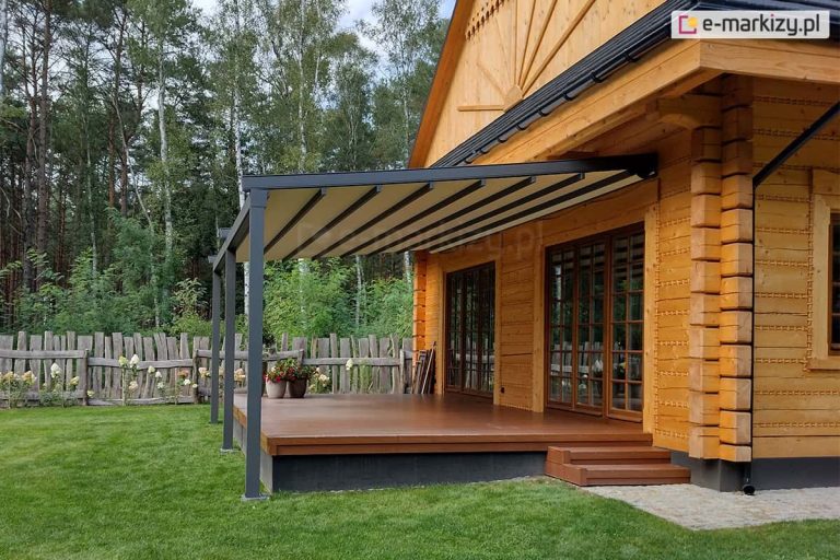 Pergola tarasowa Square o konstrukcji aluminiowej w kolorze ciemnego antracytu, zamontowana do elewacji budynku wykonanego z jasnych bali drewnianych o tradycyjnym charakterze. Zadaszenie pergoli składa się z kremowego poszycia tkaninowego rozpiętego na prowadnicach, które jest lekko nachylone pod kątem, aby zapewnić swobodny odpływ wody opadowej. Konstrukcja wspiera się na trzech pionowych, smukłych słupach o kwadratowym przekroju, które są zakotwiczone w podłożu przy krawędzi tarasu. Sam taras jest podwyższony, wykonany z gładkich desek w kolorze ciemnego brązu, a do jego wejścia prowadzą dwa niewielkie, drewniane stopnie przy ścianie po prawej stronie. Na tarasie, przy ścianie domu, widoczne są duże przeszklenia drzwi tarasowych ze szprosami. W narożniku tarasu ustawiono dwie donice z roślinami o czerwonych kwiatach. Budynek posiada charakterystyczne zdobienia snycerskie na szczycie dachu oraz czarne rynny. Tło zdjęcia stanowi gęsty las z wysokimi, zielonymi drzewami liściastymi i iglastymi, a cały teren wokół tarasu pokryty jest zadbanym trawnikiem. Po lewej stronie widoczne jest niskie, rustykalne ogrodzenie wykonane z pionowych, drewnianych sztachet, przy którym rosną jasne kwiaty ogrodowe.