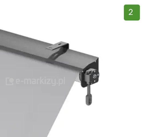 Zdjęcie prezentuje szczegółowy widok bocznego mechanizmu markizy balkonowej sklepowej umieszczonej w kasecie ochronnej. Na górze konstrukcji widoczna jest ciemnoszara, metalowa obudowa o półokrągłym profilu, która osłania rurę nawojową z tkaniną. Od góry kasety odchodzi płaski, metalowy uchwyt montażowy w kształcie litery L z podłużnym otworem na śrubę, skierowany do tyłu urządzenia. Jasnoszary materiał markizy wysuwa się z dolnej części kasety i opada ukośnie w dół. Prawy bok kasety jest zamknięty czarną maskownicą, do której przymocowany jest mechanizm sterujący. Z mechanizmu tego zwisa pionowo w dół krótki, czarny element z zaoblonym zakończeniem, służący do zaczepienia korby sterowania ręcznego. Całość przedstawiona jest na czystym, jasnym tle. W prawym górnym rogu obrazu znajduje się zielony, kwadratowy piktogram z białą cyfrą dwa.