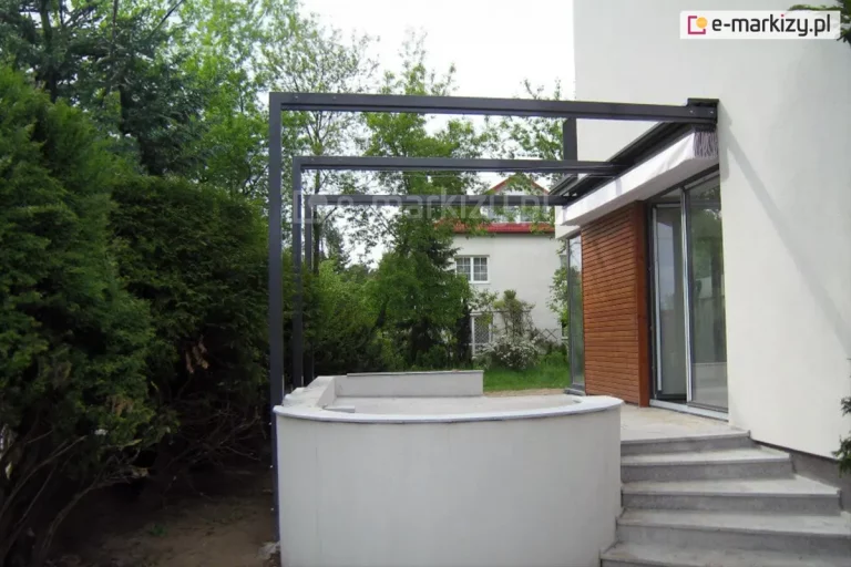 Zdjęcie prezentuje antracytową, aluminiową konstrukcję pergoli zamontowaną nad wejściem do budynku, stanowiącą nowoczesne zadaszenie tarasu ze schodami. Konstrukcja składa się z trzech smukłych, pionowych słupów połączonych z poziomymi belkami, które są zakotwiczone bezpośrednio w jasnej elewacji domu. Nad drzwiami tarasowymi widoczny jest całkowicie zwinięty pakiet białej tkaniny systemu Supro, zabezpieczony pod wąskim daszkiem ochronnym, co pozwala na pełne doświetlenie wnętrza budynku. Strefa wejściowa obejmuje betonowy podest o półkolistym kształcie oraz szerokie schody wyłożone jasnym kamieniem lub gresem. Część elewacji przy przesuwnych drzwiach przeszklonych została wykończona pionowymi panelami drewnianymi w ciepłym odcieniu. W tle widoczna jest gęsta roślinność ogrodowa z wysokimi drzewami liściastymi oraz fragment sąsiedniego budynku, co podkreśla miejski, a jednocześnie zielony charakter otoczenia.