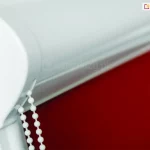 Biała kasetka rolety materiałowej na okno z ukrytym w niej mechanizmem łańcuszkowego sterowania osłoną wtapia się w kolor ramy okna, a czerwona tkanina nadaje całości wyrazistego charakteru.