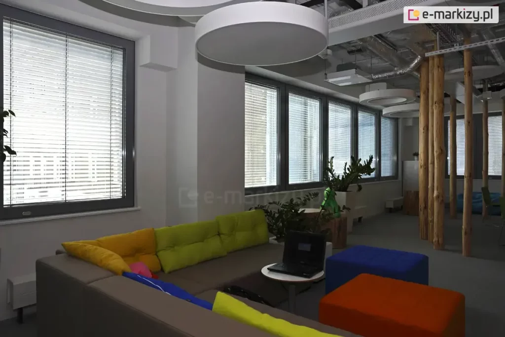Widok na nowoczesną strefę chillout w biurze typu open space, wyposażoną w system wewnętrznych żaluzji poziomych C50 w kolorze srebrnym. Żaluzje zamontowane są na szeregu okien o ciemnoszarych profilach, a ich lamele ustawione są w pozycji otwartej, co pozwala na rozproszenie światła dziennego przy zachowaniu widoczności na zewnątrz. Wnętrze charakteryzuje się niekonwencjonalnym designem: po prawej stronie widoczne są naturalne, drewniane pnie pełniące funkcję dekoracyjnych kolumn konstrukcyjnych, a na suficie zamontowano duże, okrągłe oprawy oświetleniowe oraz odkryte instalacje techniczne. Wyposażenie obejmuje narożną sofę w kolorze taupe z kolorowymi poduszkami (żółte, zielone, różowe), stolik kawowy z laptopem oraz kolorowe pufy (niebieska, pomarańczowa). Całość uzupełniają rośliny doniczkowe ustawione w białych osłonkach na parapetach oraz przy elementach drewnianych.
