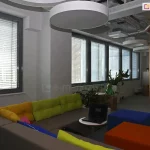 Widok na nowoczesną strefę chillout w biurze typu open space, wyposażoną w system wewnętrznych żaluzji poziomych C50 w kolorze srebrnym. Żaluzje zamontowane są na szeregu okien o ciemnoszarych profilach, a ich lamele ustawione są w pozycji otwartej, co pozwala na rozproszenie światła dziennego przy zachowaniu widoczności na zewnątrz. Wnętrze charakteryzuje się niekonwencjonalnym designem: po prawej stronie widoczne są naturalne, drewniane pnie pełniące funkcję dekoracyjnych kolumn konstrukcyjnych, a na suficie zamontowano duże, okrągłe oprawy oświetleniowe oraz odkryte instalacje techniczne. Wyposażenie obejmuje narożną sofę w kolorze taupe z kolorowymi poduszkami (żółte, zielone, różowe), stolik kawowy z laptopem oraz kolorowe pufy (niebieska, pomarańczowa). Całość uzupełniają rośliny doniczkowe ustawione w białych osłonkach na parapetach oraz przy elementach drewnianych.