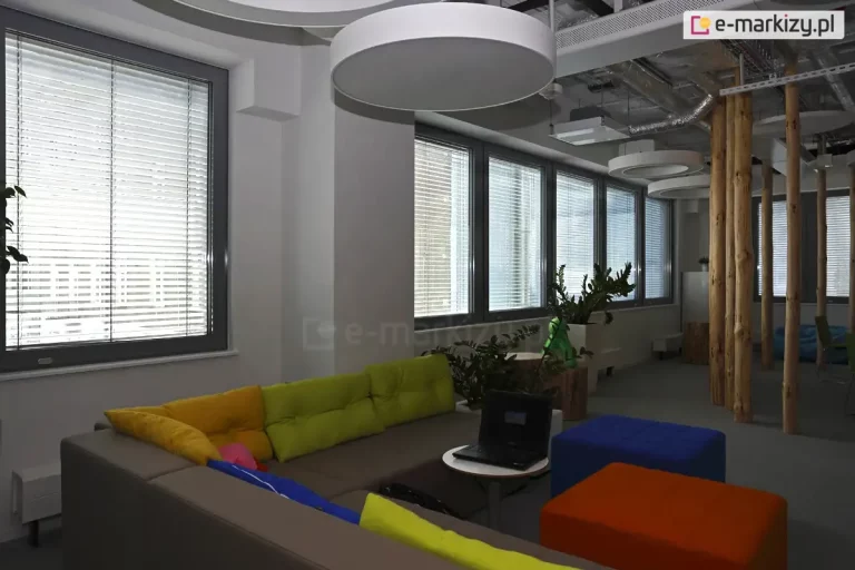 Widok na nowoczesną strefę chillout w biurze typu open space, wyposażoną w system wewnętrznych żaluzji poziomych C50 w kolorze srebrnym. Żaluzje zamontowane są na szeregu okien o ciemnoszarych profilach, a ich lamele ustawione są w pozycji otwartej, co pozwala na rozproszenie światła dziennego przy zachowaniu widoczności na zewnątrz. Wnętrze charakteryzuje się niekonwencjonalnym designem: po prawej stronie widoczne są naturalne, drewniane pnie pełniące funkcję dekoracyjnych kolumn konstrukcyjnych, a na suficie zamontowano duże, okrągłe oprawy oświetleniowe oraz odkryte instalacje techniczne. Wyposażenie obejmuje narożną sofę w kolorze taupe z kolorowymi poduszkami (żółte, zielone, różowe), stolik kawowy z laptopem oraz kolorowe pufy (niebieska, pomarańczowa). Całość uzupełniają rośliny doniczkowe ustawione w białych osłonkach na parapetach oraz przy elementach drewnianych.