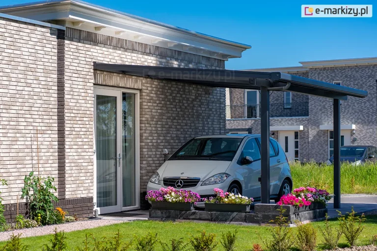 Zdjęcie prezentuje przyścienną wiatę garażową Nebbiolo marki Deponti, pełniącą funkcję zadaszenia dla miejsca postojowego. Konstrukcja z matowego aluminium w kolorze antracytowym jest zamontowana do elewacji budynku wykonanej z jasnej, cieniowanej cegły klinkierowej. Pod jednospadowym dachem z ciemnym pokryciem zaparkowany jest srebrny samochód osobowy marki Mercedes klasy A, ustawiony przodem do obiektywu. Zadaszenie wspiera się na pionowych słupach o kwadratowym przekroju, przy których znajduje się niskie obmurowanie z ciemnych bloczków, zwieńczone podłużnymi donicami z kwitnącymi różowymi i fioletowymi kwiatami. Obok wiaty widoczne są przeszklone drzwi tarasowe w białych ramkach. W tle znajdują się inne nowoczesne budynki mieszkalne o podobnej architekturze, zielony trawnik oraz roślinność ogrodowa pod bezchmurnym, błękitnym niebem.