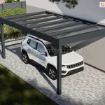 Wiata nad samochód przymocowana do ściany budynku o jasnej fasadzie. Antracytowa płaska konstrukcja i dach ze szkła chronią stojący pod nim biały pojazd przed warunkami atmosferycznymi. Przestrzeń pod zadaszeniem wyłożono kostką brukową. W tle widoczny ogród z roślinami zielonym i trawnikiem.