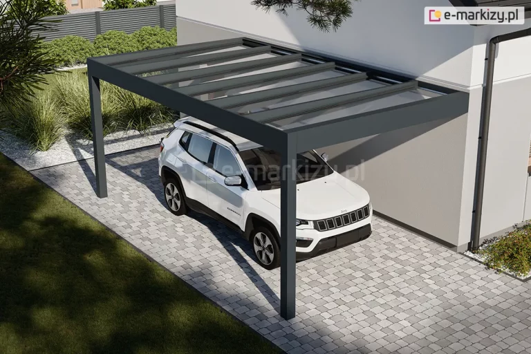 Wiata nad samochód przymocowana do ściany budynku o jasnej fasadzie. Antracytowa płaska konstrukcja i dach ze szkła chronią stojący pod nim biały pojazd przed warunkami atmosferycznymi. Przestrzeń pod zadaszeniem wyłożono kostką brukową. W tle widoczny ogród z roślinami zielonym i trawnikiem.