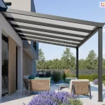 Nowoczesna wiata tarasowa Lunaro w kolorze antracytowym, zamontowana do jasnej elewacji nowoczesnego domu. Jednospadowy dach konstrukcji wypełniają panele z poliwęglanu komorowego w kolorze mlecznym, które opierają się na systemowych, ciemnych krokwiach. Pod zadaszeniem, na tarasie wyłożonym białymi płytami, znajduje się strefa wypoczynkowa z wygodnymi, beżowymi fotelami i sofą ozdobioną turkusowymi poduszkami. Aranżację dopełniają lawendowe kępy kwiatów na pierwszym planie oraz widok na ogród z zielonymi drzewami i niskim żywopłotem pod czystym niebem.