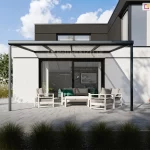 Zdjęcie prezentuje wizualizację nowoczesnego tarasu przy budynku o minimalistycznej architekturze, osłoniętego zadaszeniem Nebbiolo marki Deponti. Konstrukcja zadaszenia opiera się na antracytowym, aluminiowym stelażu z dachem wypełnionym transparentnym poliwęglanem komorowym, który rzuca liniowy cień na jasną elewację domu. Pod zadaszeniem ustawiono komplet nowoczesnych mebli ogrodowych w kolorze białym z szarymi poduszkami, składający się z sofy, dwóch foteli oraz niskiego stolika kawowego. Szerokie, przeszkolone drzwi przesuwne prowadzą z tarasu bezpośrednio do wnętrza salonu. Nawierzchnia tarasu wykonana jest z dużych, jasnoszarych płyt betonowych, a w jego otoczeniu znajdują się ozdobne trawy w donicach oraz niska roślinność ogrodowa. Całość kompozycji utrzymana jest w chłodnej, nowoczesnej stylistyce pod jasnym niebem z delikatnymi chmurami.