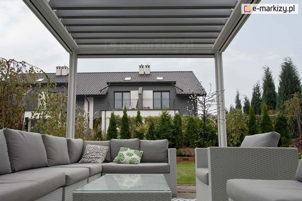 Widok z wnętrza pergoli tarasowej SB400 Seltna strefę wypoczynkową i ogród. Konstrukcja pergoli w jasnym kolorze białym posiada dach z ruchomych, równolegle ułożonych lameli. Pod zadaszeniem znajduje się zestaw mebli ogrodowych wykonanych z szarego technorattanu, składający się z dużej narożnej sofy z grubymi, szarymi poduszkami, dopasowanych stylistycznie dwóch foteli oraz niskiego stolika kawowego ze szklanym blatem. Na sofie widoczne są dwie poduszki dekoracyjne: jedna z motywem liści monstery, druga z geometrycznym wzorem. W tle, za rzędem gęstych tui, widoczny jest nowoczesny dom piętrowy o szarej elewacji z ciemnym, spadzistym dachem i balkonami. Niebo jest zachmurzone, a cała aranżacja prezentuje nowoczesny design przestrzeni przydomowej.