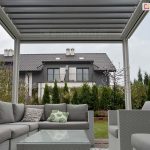 Widok z wnętrza pergoli tarasowej SB400 Seltna strefę wypoczynkową i ogród. Konstrukcja pergoli w jasnym kolorze białym posiada dach z ruchomych, równolegle ułożonych lameli. Pod zadaszeniem znajduje się zestaw mebli ogrodowych wykonanych z szarego technorattanu, składający się z dużej narożnej sofy z grubymi, szarymi poduszkami, dopasowanych stylistycznie dwóch foteli oraz niskiego stolika kawowego ze szklanym blatem. Na sofie widoczne są dwie poduszki dekoracyjne: jedna z motywem liści monstery, druga z geometrycznym wzorem. W tle, za rzędem gęstych tui, widoczny jest nowoczesny dom piętrowy o szarej elewacji z ciemnym, spadzistym dachem i balkonami. Niebo jest zachmurzone, a cała aranżacja prezentuje nowoczesny design przestrzeni przydomowej.