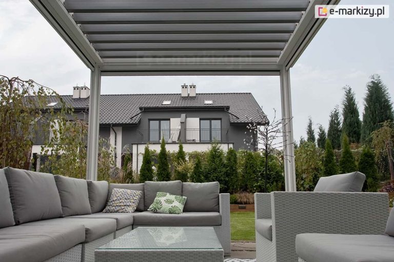Widok z wnętrza pergoli tarasowej SB400 Seltna strefę wypoczynkową i ogród. Konstrukcja pergoli w jasnym kolorze białym posiada dach z ruchomych, równolegle ułożonych lameli. Pod zadaszeniem znajduje się zestaw mebli ogrodowych wykonanych z szarego technorattanu, składający się z dużej narożnej sofy z grubymi, szarymi poduszkami, dopasowanych stylistycznie dwóch foteli oraz niskiego stolika kawowego ze szklanym blatem. Na sofie widoczne są dwie poduszki dekoracyjne: jedna z motywem liści monstery, druga z geometrycznym wzorem. W tle, za rzędem gęstych tui, widoczny jest nowoczesny dom piętrowy o szarej elewacji z ciemnym, spadzistym dachem i balkonami. Niebo jest zachmurzone, a cała aranżacja prezentuje nowoczesny design przestrzeni przydomowej.