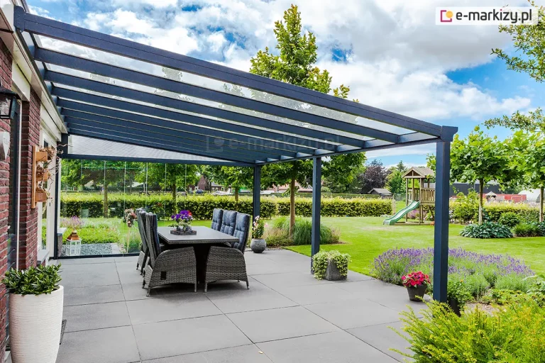 Zdjęcie ukazuje perspektywę szerokiego zadaszenia tarasu Terano z wypełnieniem szklanym, zamontowanego do budynku z czerwonej cegły. Konstrukcja antracytowa składa się z licznych krokwi dachowych oraz systemu wsporczego opartego na słupach o przekroju kwadratowym. Pod zadaszeniem ustawiono duży stół obiadowy z sześcioma wysokimi fotelami technorattanowymi w kolorze szarym. Taras wyłożony jest wielkoformatowymi, szarymi płytami. W tle widoczny jest rozległy ogród z placem zabaw dla dzieci (huśtawka i zjeżdżalnia), niskimi żywopłotami, fioletową lawendą oraz drzewami pod dynamicznym, zachmurzonym niebem.