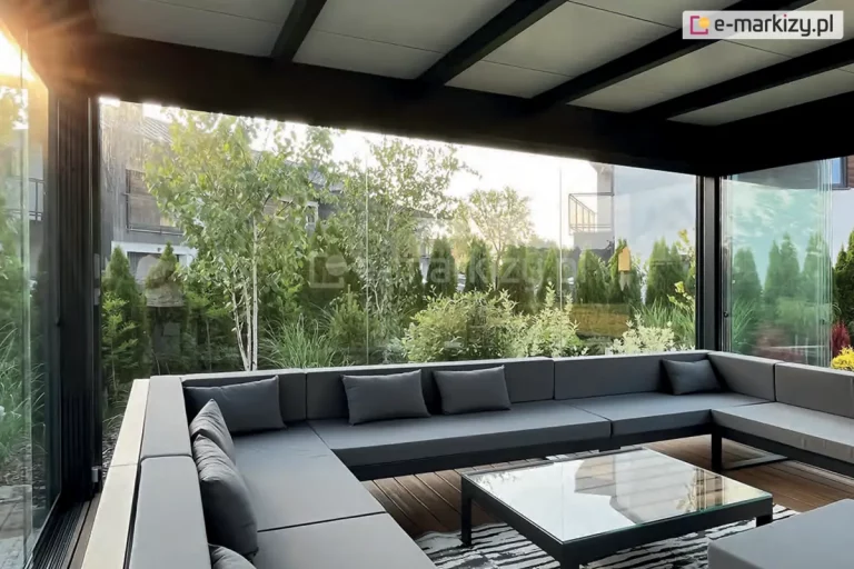 Przestrzeń wypoczynkowa w szklanej pergoli z widokiem na ogród. Wnętrze wyposażone w duży, narożny zestaw mebli ogrodowych z szarej tapicerki, złożony z prostych segmentów z oparciami i poduszkami. Na środku znajduje się niski stolik z blatem ze szkła i czarną ramą. Pod nim dywan w czarno-białe pasy. Ściany pergoli wykonane z dużych tafli szkła bez ram - system przesuwny Miros, przez które widoczny jest zadbany ogród z mieszanką roślin liściastych i iglastych oraz częściowo widoczne budynki. Promienie słońca wlewają się do wnętrza od lewej strony zdjęcia.