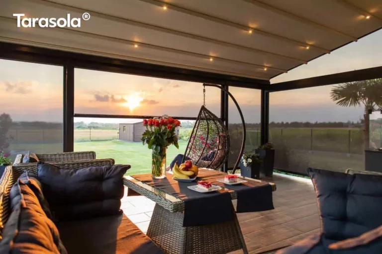 Wnętrze luksusowo urządzonego tarasu pod zadaszeniem tkaninowym Elegancy Tarasola, uchwycone w ciepłym świetle zachodzącego słońca. Pergola wyposażona jest w jasny, rozsuwany dach z wbudowanym punktowym oświetleniem LED, które nadaje przestrzeni przytulny charakter. Boki konstrukcji zostały osłonięte transparentnymi refleksolami Zip, chroniącymi przed wiatrem, nie ograniczając jednocześnie widoku na rozległy ogród i horyzont. Na tarasie znajduje się zestaw wypoczynkowy z technorattanu z ciemnymi poduszkami oraz stół nakryty bieżnikami, na którym stoją owoce, desery i wazon z czerwonymi różami. W tle widoczny jest wiszący fotel typu kokon oraz nastrojowy krajobraz z polami, oświetlony niskim słońcem przebijającym się przez chmury.