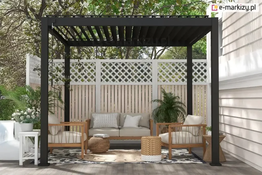 Nowoczesna pergola tarasowa Gloria w kolorze antracytowym z otwartym dachem lamelowym, zamontowana na drewnianym podeście przy jasnej elewacji budynku. Konstrukcja opiera się na ciemnych, matowych profilach aluminiowych, a dach tworzą równolegle ustawione czarne lamele przepuszczające światło słoneczne. Wewnątrz znajduje się strefa wypoczynkowa z drewnianą sofą i dwoma fotelami o jasnej tapicerce, ozdobionymi poduszkami. Pod meblami leży wzorzysty dywan zewnętrzny oraz okrągła pleciona mata. Dodatki stanowią plecione pufy, stoliki pomocnicze z drewna i wikliny oraz liczne rośliny doniczkowe, w tym palmy i kwiaty w białych donicach. Tło pergoli stanowi białe ogrodzenie z ozdobną kratownicą w górnej części, za którą widoczne są gęste korony drzew. Z lewej strony górnej ramy zwisa metalowy drążek korbowy do sterowania zadaszeniem.