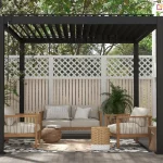 Nowoczesna pergola tarasowa Gloria w kolorze antracytowym z otwartym dachem lamelowym, zamontowana na drewnianym podeście przy jasnej elewacji budynku. Konstrukcja opiera się na ciemnych, matowych profilach aluminiowych, a dach tworzą równolegle ustawione czarne lamele przepuszczające światło słoneczne. Wewnątrz znajduje się strefa wypoczynkowa z drewnianą sofą i dwoma fotelami o jasnej tapicerce, ozdobionymi poduszkami. Pod meblami leży wzorzysty dywan zewnętrzny oraz okrągła pleciona mata. Dodatki stanowią plecione pufy, stoliki pomocnicze z drewna i wikliny oraz liczne rośliny doniczkowe, w tym palmy i kwiaty w białych donicach. Tło pergoli stanowi białe ogrodzenie z ozdobną kratownicą w górnej części, za którą widoczne są gęste korony drzew. Z lewej strony górnej ramy zwisa metalowy drążek korbowy do sterowania zadaszeniem.
