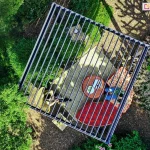 Widok z lotu ptaka na nowoczesną, kwadratową pergolę bioklimatyczną Gloria w ciemnym kolorze, umieszczoną w gęsto porośniętym roślinami ogrodzie. Dach konstrukcji składa się z równoległych, aluminiowych lamel ustawionych w pozycji półotwartej, które rzucają liniowe cienie na strefę wypoczynkową poniżej. Pod pergolą znajduje się jasny, kwadratowy dywan zewnętrzny, na którym ustawiono meble ogrodowe: okrągły stolik z dekoracyjnym blatem oraz narożną sofę z czerwonymi poduszkami w jasne, roślinne wzory. Całość otoczona jest bogatą roślinnością, krzewami i drzewami, a podłoże wokół dywanu wysypane jest ciemnym żwirem lub korą. Na lamelach dachu widoczne są odbicia światła słonecznego, podkreślające metaliczną strukturę konstrukcji.