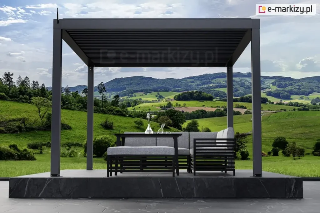Antracytowa pergola aluminiowa SB350 Aluprof z zamkniętym dachem lamelowym i automatyką bezprzewodową Blebox nad podestem z marmuru, taras z widokiem na góry oraz zielone wzgórza z roślinnością. Na podeście ustawiono narożnik oraz siedzisko z wygodnymi poduchami w szarym kolorze oraz stolik ze szklanym wazonem z kilkoma kwiatkami.