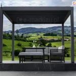 Antracytowa pergola aluminiowa SB350 Aluprof z zamkniętym dachem lamelowym i automatyką bezprzewodową Blebox nad podestem z marmuru, taras z widokiem na góry oraz zielone wzgórza z roślinnością. Na podeście ustawiono narożnik oraz siedzisko z wygodnymi poduchami w szarym kolorze oraz stolik ze szklanym wazonem z kilkoma kwiatkami.