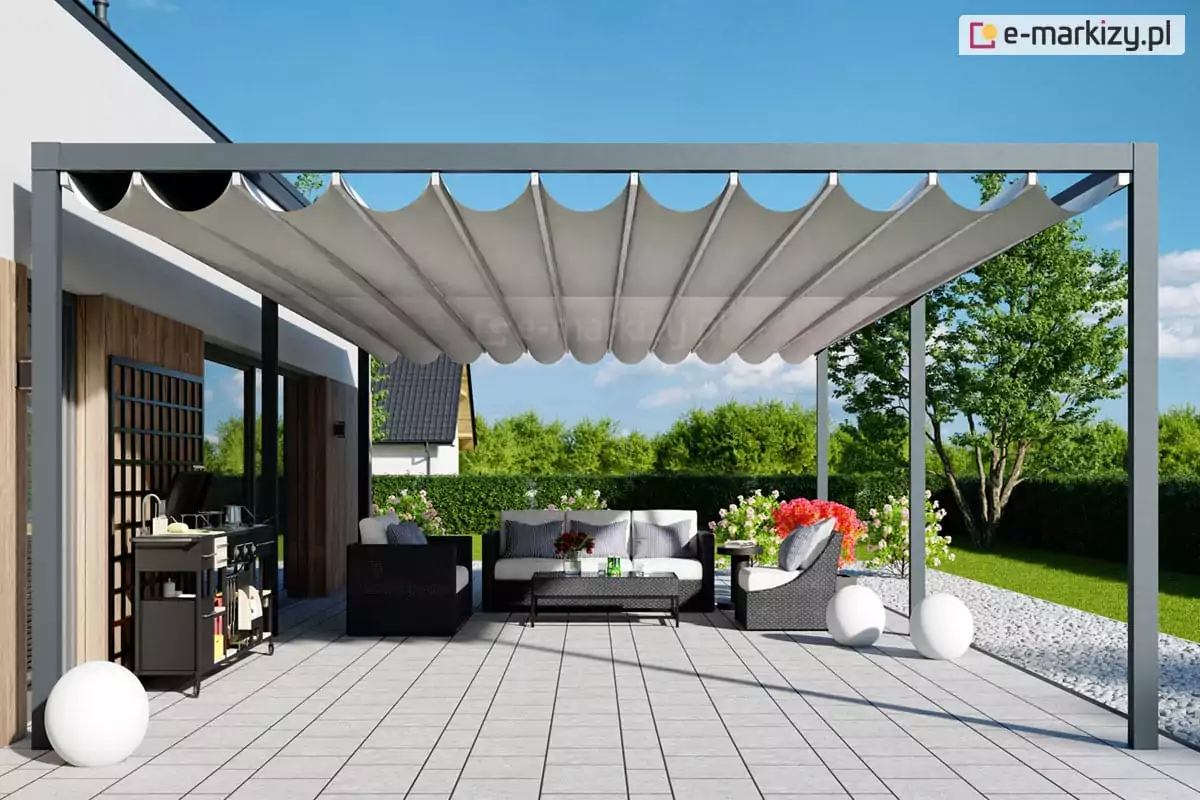 Zdjęcie przedstawia wolnostojącą pergolę tarasową o nowoczesnej, minimalistycznej konstrukcji wykonanej z profili aluminiowych w kolorze ciemnoszarym. Zadaszenie pergoli stanowi jasny, biały materiał ułożony w głębokie, regularne fale, co jest charakterystyczne dla systemów rzymskich (tkaninowych). Konstrukcja wspiera się na smukłych, pionowych słupach i jest ustawiona na przestronnym tarasie wyłożonym dużymi, jasnoszarymi płytami betonowymi, który otoczony jest pasem białego żwiru dekoracyjnego. Pod pergolą zaaranżowano strefę wypoczynkową z zestawem mebli ogrodowych z technorattanu, składającym się z sofy i foteli z jasnymi poduchami. Obok mebli znajduje się nowoczesny, czarny grill ogrodowy na kółkach oraz dekoracyjne oświetlenie w formie dużych, białych kul świetlnych leżących bezpośrednio na posadzce. W tle widoczny jest zadbany ogród z zielonym trawnikiem, żywopłotem oraz dom jednorodzinny z elewacją częściowo wykończoną pionowymi lamelami drewnianymi.