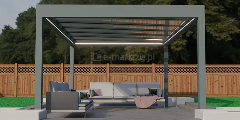 Wolnostojąca, nowoczesna konstrukcja zadaszenia tarasowego Spekta Glass, zintegrowana z otwartą przestrzenią ogrodu. Konstrukcja nośna została wykonana z grubych profili aluminiowych w kolorze antracytowym, które tworzą geometryczną, prostopadłościenną ramę wspartą na czterech solidnych słupach. Dach konstrukcji jest w całości przeszklony, co pozwala na maksymalne doświetlenie wnętrza światłem dziennym, a dodatkowo wyposażony w zintegrowane liniowe oświetlenie LED zamontowane wzdłuż profili dachu. Wewnątrz zadaszonej strefy zaaranżowano miejsce do wypoczynku, wyposażone w komplet mebli tarasowych: narożną sofę z jasnoszarymi, miękkimi poduchami oraz dekoracyjnymi kocami. Podłoże pod zabudową stanowią wielkoformatowe płyty tarasowe w odcieniach szarości, które tworzą stabilną i estetyczną bazę. W narożnikach konstrukcji widoczne są niskie, białe donice o geometrycznych kształtach, pełniące funkcję architektonicznego obramowania tarasu. Całość kompozycji dopełnia wysokie drewniane ogrodzenie z ażurowym wykończeniem u góry oraz ściana gęstej, zielonej roślinności w tle, co podkreśla spójność nowoczesnej architektury z otaczającą naturą.