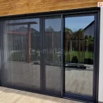 Duże okno z wyjściem na taras z płytek o kolorze drewna, przesłonięte częściowo dopasowaną kolorystycznie do ramy okna moskitierą plisowaną Top Zag z szarą siatką. Moskitiera zamontowana wewnątrz wnęki na ścianie o jasnej elewacji.