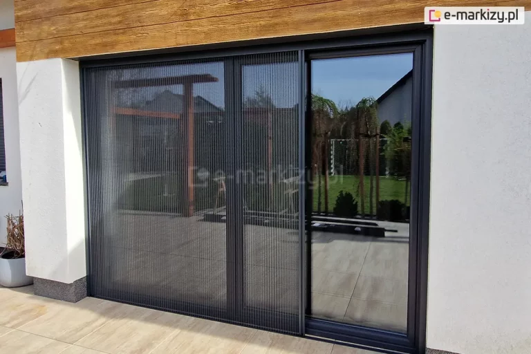 Duże okno z wyjściem na taras z płytek o kolorze drewna, przesłonięte częściowo dopasowaną kolorystycznie do ramy okna moskitierą plisowaną Top Zag z szarą siatką. Moskitiera zamontowana wewnątrz wnęki na ścianie o jasnej elewacji.