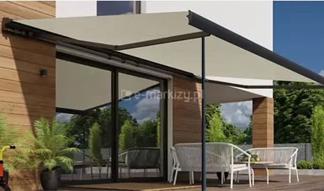 Taras przy budynku mieszkalnym, nad którym rozłożona jest markiza w jasnym, beżowym kolorze. Standardowa konstrukcja markizy Palladio Selt z napędem ręcznym wzbogacona została o pionowe podpory teleskopowe w kolorze grafitowym, które są przytwierdzone do podłoża. Ich rolą jest wsparcie belki przedniej i ochrona przed falowaniem podczas podmuchów wiatru. Osłona jest zamontowana na elewacji łączącej biały tynk z pionowymi panelami o wyglądzie jasnego drewna. Pod markizą znajduje się zestaw mebli wypoczynkowych, w tym białe fotele z plecionki oraz stolik, ustawione na ciemnym, drewnianym tarasie. W dużych, szklanych drzwiach przesuwnych odbija się błękitne niebo z białymi chmurami oraz korony drzew. Po lewej stronie widoczna jest zielona roślina w czarnej donicy, a w tle widać nowoczesne ogrodzenie i fragmenty zadbanej roślinności ogrodowej.