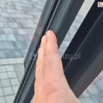 Zdjęcie prezentuje zbliżenie na pionowy profil krawędziowy aluminiowych drzwi szklanych systemu Sotro, pełniący funkcję zintegrowanego pochwytu. Profil w matowym kolorze antracytowym posiada specjalnie wyprofilowany, ergonomiczny kształt z zaokrąglonym grzbietem, który umożliwia wygodne przesuwanie skrzydła dłonią na dowolnej wysokości, co ilustruje oparta o niego ręka. Perspektywa ujęcia od góry ukazuje w tle fragment tarasu wyłożonego szarą kostką brukową, podkreślając nowoczesny charakter wykończenia detali konstrukcyjnych.