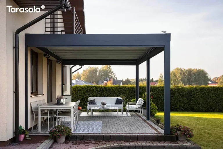 Nowoczesna, przyścienna pergola tarasowa essential Tarasola w kolorze ciemnoantracytowym, zamontowana nad tarasem z kostki brukowej przy domu jednorodzinnym. Konstrukcja bioklimatyczna posiada dach z ruchomych lameli oraz otwarte ściany boczne, co zapewnia swobodny przepływ powietrza i widok na ogród. Pod zadaszeniem wydzielono dwie strefy: jadalnianą z szarym stołem i krzesłami ustawionymi na wzorzystym dywanie zewnętrznym oraz wypoczynkową z białą sofą, fotelami i stolikiem kawowym. Na elewacji budynku, pod konstrukcją, widoczne są zamontowane promienniki ciepła oraz punkty świetlne. W tle znajduje się niski żywopłot, trawnik oraz inne zabudowania jednorodzinne pod jasnym niebem w ciągu dnia. Scena prezentuje funkcjonalne wykorzystanie aluminiowej pergoli do stworzenia komfortowego salonu na świeżym powietrzu, zintegrowanego z bryłą budynku i otaczającą go zielenią.