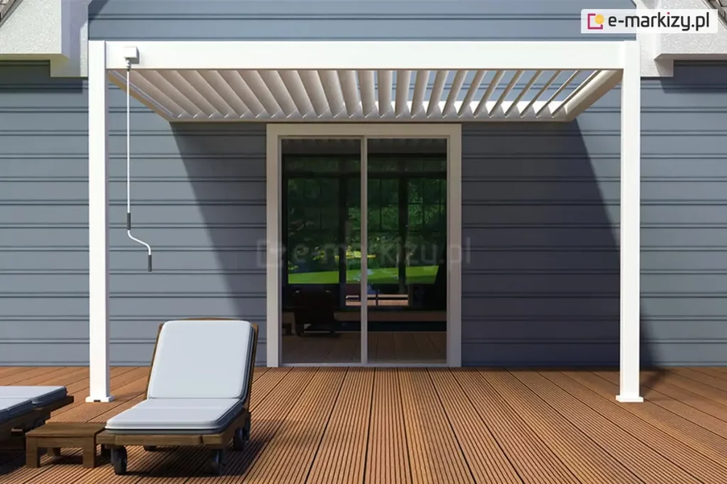 Nowoczesna biała pergola lamelowa Marlo zamontowana na tarasie przy budynku mieszkalnym. Konstrukcja pergoli opiera się na dwóch pionowych, kwadratowych słupach z przodu oraz jest przytwierdzona bezpośrednio do elewacji budynku, która wykończona jest poziomym sidingiem w kolorze szarym. Zadaszenie składa się z ruchomych, równolegle ułożonych białych lameli, które są obecnie ustawione pod kątem, pozwalając na częściowy dopływ światła słonecznego. Po lewej stronie konstrukcji widoczna jest zwisająca biała korba służąca do ręcznej regulacji kąta nachylenia lameli. Pod pergolą znajduje się taras wykonany z desek kompozytowych lub drewnianych w naturalnym, miodowym odcieniu, ułożonych prostopadle do elewacji. Na tarasie ustawiony jest ciemnobrązowy leżak z jasnoszarą poduszką oraz mały, niski stolik kawowy. W tle widać duże, przeszklone drzwi przesuwne prowadzące do wnętrza domu, w których odbija się zieleń ogrodu. Cień rzucany przez pergolę na elewację i podłogę tarasu jest wyraźny, co wskazuje na słoneczną pogodę.